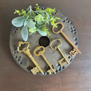 May include: An arrangement of vintage keys in brass and copper tones, displayed on a dark, circular metal plate with a central hole and numerous smaller holes. Greenery adds a touch of nature. The keys vary in design, showcasing intricate details.