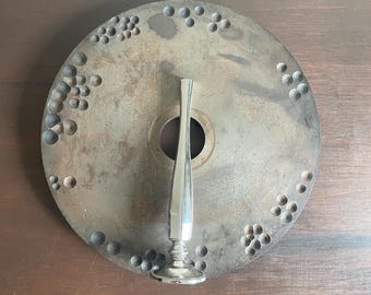 May include: A silver metal bottle stopper with a round base and a long, thin neck. The stopper is sitting on top of a round, metal disc with a pattern of small holes.