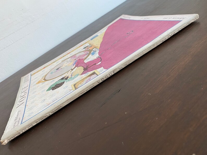 Puede incluir: Una edici&oacute;n vintage de la revista Vogue del 15 de febrero de 1918 con una colorida ilustraci&oacute;n de una persona con un vestido largo magenta mirando en un espejo. La portada incluye el texto Vogue, New York City y Spring Millinery Number.