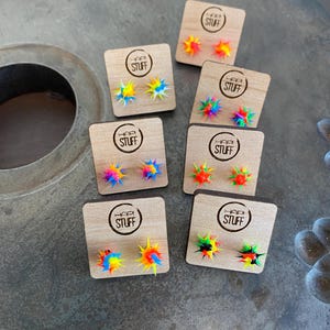 May include: Six pairs of colorful, spiky earrings displayed on small wooden squares. Each square has the text "Happy Stuff" printed on it.