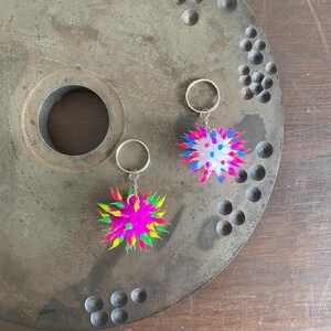 May include: Two colorful, spiky keychains. One is pink with yellow and green accents, the other is white with blue and pink accents. Both have silver key rings. The keychains are on a textured, gray metal surface.
