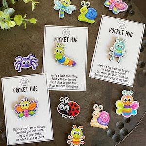 May include: A collection of colorful wooden character pins featuring insects and creatures like butterflies, snails, ladybugs, and caterpillars. Three pins are attached to white cards labeled POCKET HUG with printed messages about care and affection.