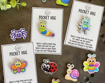 May include: A collection of colorful wooden character pins featuring insects and creatures like butterflies, snails, ladybugs, and caterpillars. Three pins are attached to white cards labeled POCKET HUG with printed messages about care and affection.