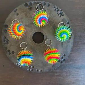 Puede incluir: Cinco llaveros coloridos, puntiagudos, con colores de arcoíris y anillos de llaves plateados. Los llaveros están hechos de un material suave y flexible y tienen forma de bola. Están dispuestos en círculo sobre una superficie gris.