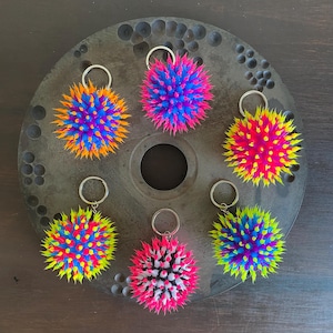 Könnte beinhalten: Sechs bunte, stachelige Stressbälle mit Schlüsselanhängern. Die Bälle bestehen aus weichem, quietschweichem Silikon und sind in verschiedenen Farben erhältlich, darunter Pink, Blau, Gelb, Grün und Orange.