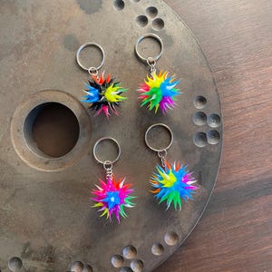 Puede incluir: Cuatro llaveros de bola con pinchos coloridos con anillos de llave plateados. Los llaveros están sobre una superficie metálica con un patrón circular.