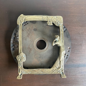 Puede incluir: Un soporte decorativo de latón con un diseño ornamentado que presenta una figura femenina y motivos florales. El soporte tiene un marco cuadrado y una base redonda.