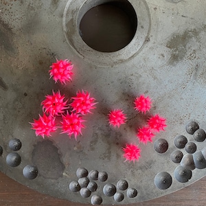 May include: A collection of bright pink, spiky, star-shaped objects scattered on a large, circular, gray metal surface. The metal surface has a large central hole and a pattern of small, dark gray spheres along the edge. The objects appear to be decorative.