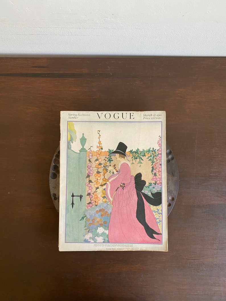 May include: A vintage Vogue magazine cover from March 15, 1924, featuring a woman in a pink dress and black hat standing in a garden setting. The cover text reads "Spring Fashions Number" and "March 15, 1924 Price 25 Cents".