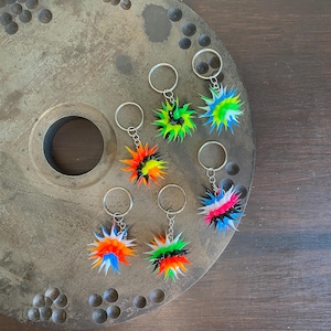 May include: Six colorful spiky keychains with silver key rings. The keychains are made of rubber and have a starburst shape. The colors include black, white, red, orange, yellow, green, and blue.