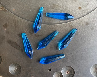 May include: Seven blue crystal teardrop-shaped pendants, with a faceted design, are scattered on a dark gray surface.