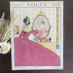 Puede incluir: Una portada de revista Vogue vintage del 15 de febrero de 1918 titulada Spring Millinery Number. La ilustraci&oacute;n muestra a una persona con un vestido magenta y un tocado verde mir&aacute;ndose en un espejo ovalado. El fondo presenta un papel tapiz floral.