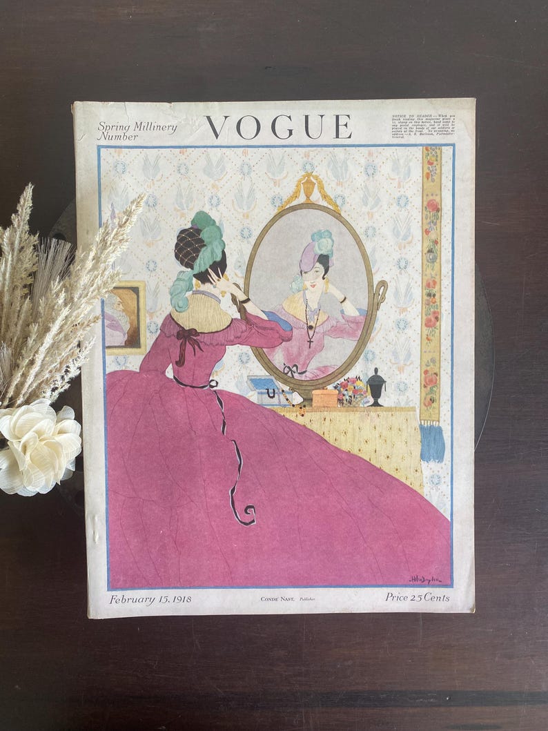 Puede incluir: Una portada de revista Vogue vintage del 15 de febrero de 1918 titulada Spring Millinery Number. La ilustraci&oacute;n muestra a una persona con un vestido magenta y un tocado verde mir&aacute;ndose en un espejo ovalado. El fondo presenta un papel tapiz floral.