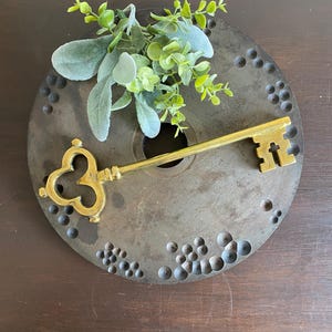 May include: A decorative brass key rests on a dark, textured metal disc, adorned with small, circular indentations. Green and gray faux foliage adds a touch of nature. The key's intricate design and the disc's rustic appearance create a vintage aesthetic.