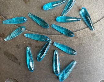 May include: A collection of 15 teal blue glass teardrop shaped beads. The beads are arranged on a dark gray surface.