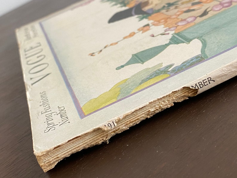 May include: A vintage Vogue magazine cover from 1917, featuring a colorful illustration of a woman in a garden setting. The cover text reads "Spring Fashions Number".