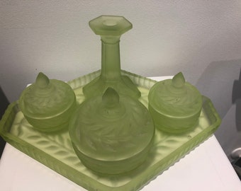 Puede incluir: Un juego de tocador de vidrio verde con una bandeja, tres frascos y un candelero alto. Los frascos y la bandeja tienen un diseño de hojas.