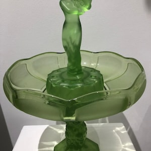 Puede incluir: Un cuenco de pedestal de vidrio verde con una figura femenina esculpida sosteniendo uvas en la parte superior. El cuenco tiene un borde festoneado y una base acanalada.