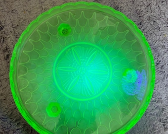 May include: A round, bright green glass cake stand with a scalloped edge. The center features a floral design, and the stand has three small feet. The glass has a glowing appearance.