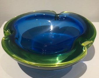 May include: A vintage ashtray with a unique design. The ashtray features a vibrant blue interior and a green rim. The glass ashtray has a rounded shape with four curved indentations. The ashtray is made of glass.