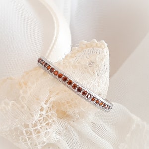 Peut inclure: Une délicate bague en argent avec une rangée de petites pierres précieuses brunes. La bague repose sur un tissu en dentelle blanche.