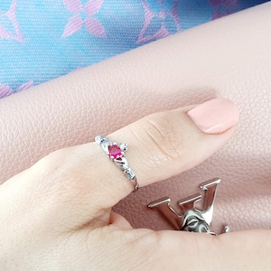 Może przedstawiać: Srebrny pierścionek z czerwonym kamieniem szlachetnym osadzonym w koronie. Pierścionek noszony jest na kciuku.