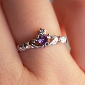 May include: A silver ring featuring a purple heart-shaped gemstone and a smaller clear gemstone on the band. The ring is a Claddagh ring, a traditional Irish design.