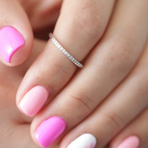 Op de afbeelding: Een fijne zilveren ring versierd met kleine, sprankelende stenen. De ring zit om een vinger met een manicure met roze en witte nagellak.