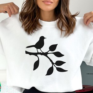 May include: White sweatshirt with a black silhouette of a bird perched on a branch with leaves.