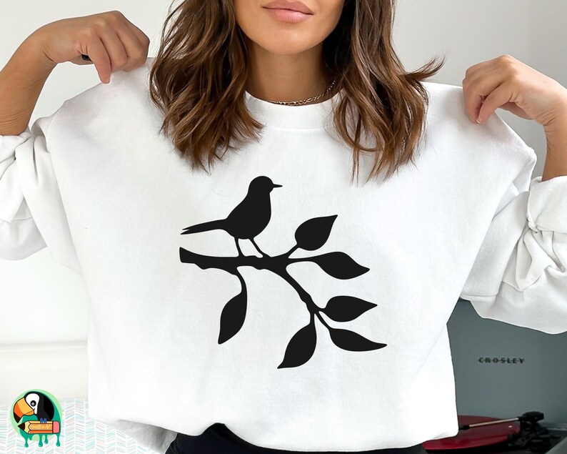 May include: White sweatshirt with a black silhouette of a bird perched on a branch with leaves.