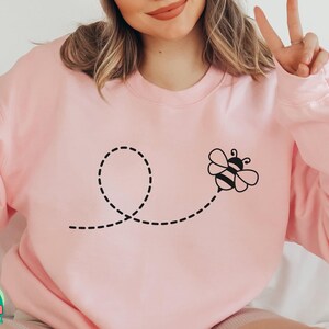 Op de afbeelding: Een roze sweatshirt met een zwarte bij en een gestippeld lijntjesontwerp. De bij vliegt in een cirkel.