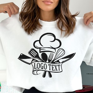 K&ouml;nnte beinhalten: Wei&szlig;es Sweatshirt mit einem schwarzen Kochhut und gekreuzten K&uuml;chenutensilien-Design. Der Text "LOGO TEXT" ist auf einem Banner unter dem Design gedruckt.
