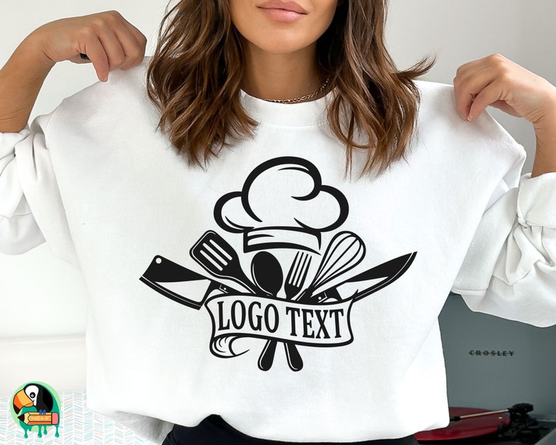K&ouml;nnte beinhalten: Wei&szlig;es Sweatshirt mit einem schwarzen Kochhut und gekreuzten K&uuml;chenutensilien-Design. Der Text "LOGO TEXT" ist auf einem Banner unter dem Design gedruckt.
