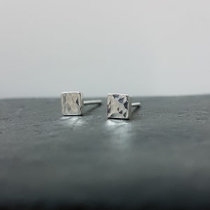 May include: A pair of small, square stud earrings. The earrings are silver-coloured with a hammered texture on the surface. The posts are thin and silver. The earrings are displayed on a dark grey surface.