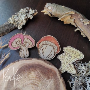 Peut inclure: Trois ornements en bois en forme de champignon. L'un est rose et blanc, l'autre est orange et blanc, et le dernier est jaune. Les ornements sont sur une surface en bois avec le mot "Korka" écrit dessus.