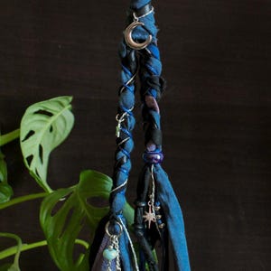 Op de afbeelding: Gevlochten haaraccessoire met blauwe en zwarte stof. Zilveren bedels, waaronder sterren en een maan, sieren het geheel. Het accessoire heeft een bohemien of heksenachtige uitstraling met een donkere achtergrond.