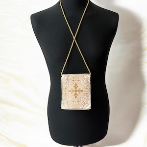May include: A small rectangular crossbody bag featuring a cream-colored fabric base with an intricate gold-toned damask pattern. The bag is suspended by a thin, matching gold-toned chain strap that forms an X-shape across the upper section.
