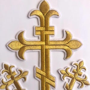 Könnte beinhalten: Drei goldene gestickte Kreuze mit weißen Umrissen. Das größte Kreuz ist ein traditionelles christliches Kreuz mit einem Fleur-de-lis-Design oben und unten. Die beiden kleineren Kreuze sind einfache Kreuze mit Fleur-de-lis-Designs an den Enden.