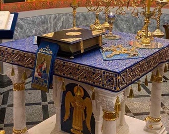 Puede incluir: Una mesa de mármol blanco con detalles dorados y un mantel con estampado azul y dorado. Un libro cerrado con una cruz dorada en la portada se encuentra sobre la mesa. La mesa está en una habitación con un mural de figuras religiosas en la pared.