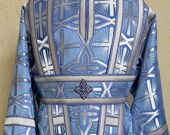 Puede incluir: Vestidura litúrgica azul claro con un detallado patrón geométrico plateado y blanco. La túnica presenta mangas largas, cuello alto y cinturón decorativo. El diseño incluye cruces y otros símbolos religiosos.