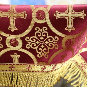May include: Burgundy fabric with gold embroidery featuring a repeating pattern of crosses and floral designs. The fabric has a gold fringe trim.