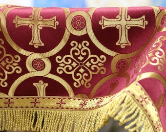 Puede incluir: Tela color burdeos con bordado dorado que presenta un patrón repetido de cruces y diseños florales. La tela tiene un ribete de flecos dorados.