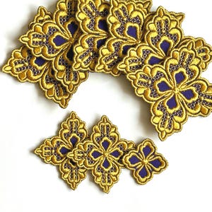 May include: Gold embroidered appliques with a floral design and a deep purple center. The appliques are arranged on a white surface. The embroidery has a detailed, ornate pattern.