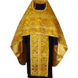 Może przedstawiać: Złota szata liturgiczna ze skomplikowanymi wzorami krzyży i kwiatów. Szata posiada duży, ozdobny kaptur i długi, dekoracyjny panel z przodu, zapinany na guziki. Tkanina ma satynowy połysk.