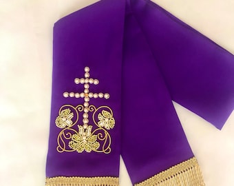 May include: A purple liturgical stole with a gold cross and floral embroidery. The cross is adorned with white pearls. Gold fringe trims the bottom edges of the stole. The stole is made of a smooth, satin-like fabric.