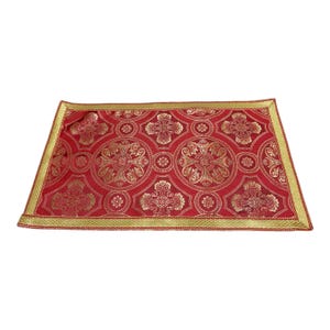 May include: A rectangular fabric mat featuring a deep red background with a repeating gold metallic pattern of circular floral motifs and crosses. The edges are finished with a wide, textured gold border that frames the intricate design.
