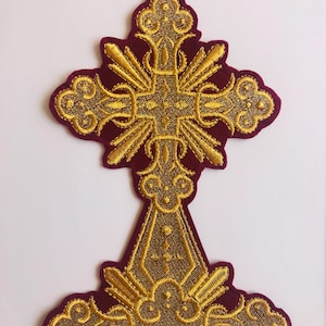 Könnte beinhalten: Ein gestickter Kreuz-Patch in Gold mit aufwendigen Details auf einem burgunderfarbenen Filzgrund. Das Kreuz ist verziert und hat eine spitze Basis.