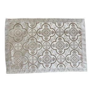 May include: A rectangular, silver decorative textile featuring a repeating pattern of crosses and floral motifs. The border has a detailed, repeating design. The fabric has a subtle sheen, giving it a luxurious appearance.