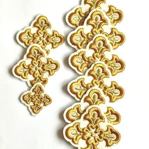 Könnte beinhalten: Eine Sammlung von gestickten Aufnähern mit einem goldenen floralen Design auf cremefarbenem Hintergrund. Die Aufnäher haben eine Vierpassform mit aufwendigen Details. Sie dienen wahrscheinlich dekorativen Zwecken, wie z. B. zum Verzieren von Kleidung oder Accessoires.