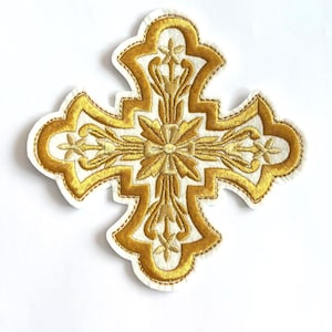 Puede incluir: Parche bordado con forma de cruz dorada con intrincados diseños florales y de hojas. La cruz está delineada con hilo dorado y tiene un fondo blanco. Ideal para ropa o proyectos de manualidades.